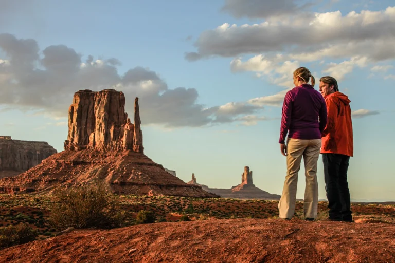 Een man en vrouw staan met de rug naar de camera en kijken uit over de rode rotsformaties van Monument Valley bij zonsondergang