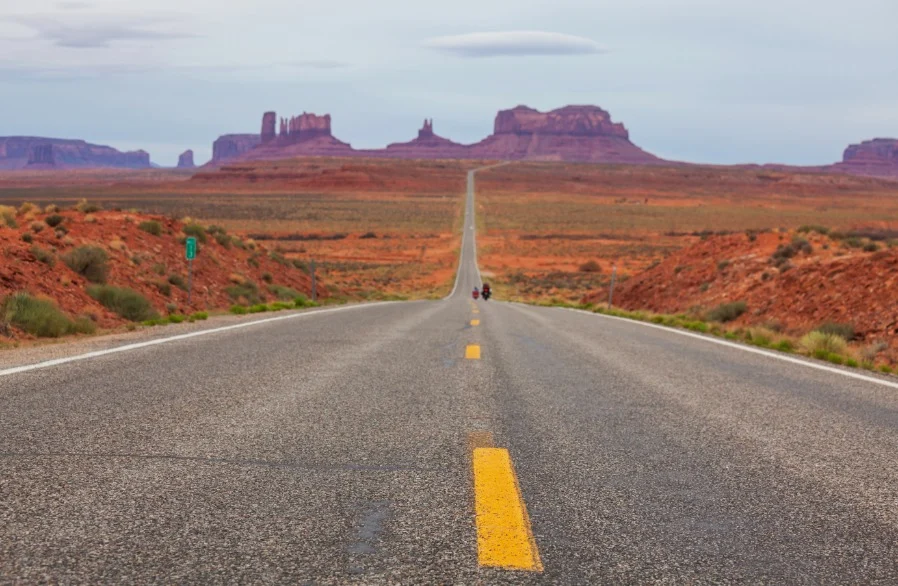 Een lange rechte asfaltweg met gele middenlijn loopt door Monument Valley naar de kenmerkende rode rotsformaties in de verte