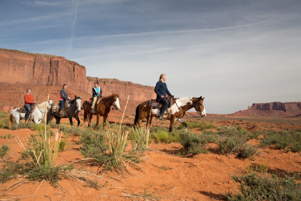 Vier ruiters te paard rijden door het rode zandlandschap van Monument Valley met rotsformaties op de achtergrond