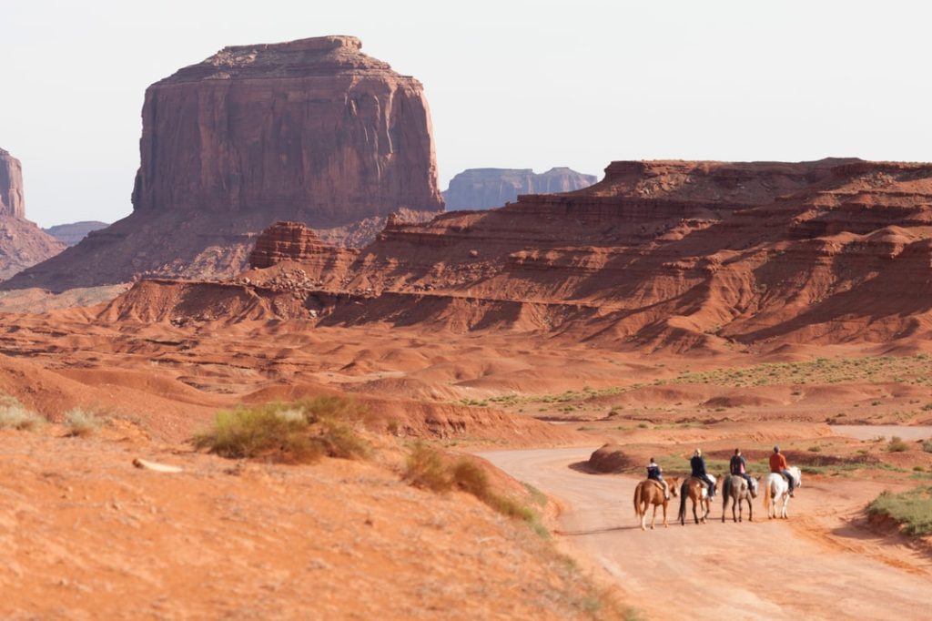 Vier ruiters te paard rijden over een zandpad tussen de roodgekleurde rotsformaties van Monument Valley