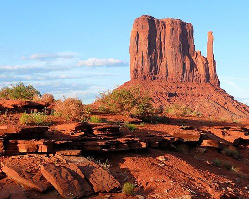 Hoge rode rotstoren met struiken en verweerde rotsplaten op de voorgrond onder een blauwe lucht in Monument Valley