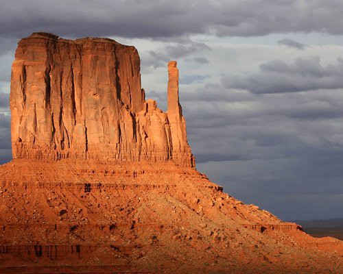Hoge rode rotsformatie met steile wanden die oplicht tegen een donkere bewolkte lucht in Monument Valley