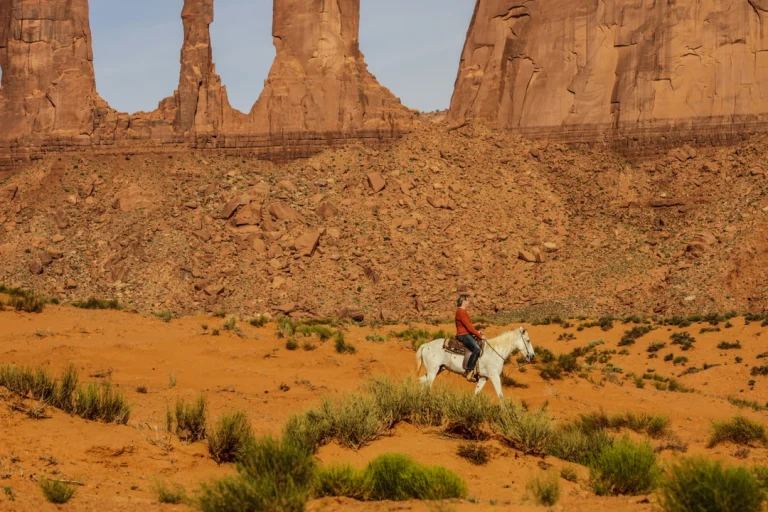Een ruiter op een wit paard rijdt door de rode woestijnvlakte van Monument Valley met hoge rotspilaren op de achtergrond