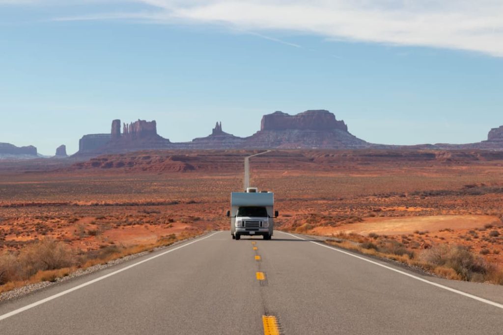Kleine camper rijdt over een kaarsrechte weg met de kenmerkende rotsformaties van Monument Valley in de verte