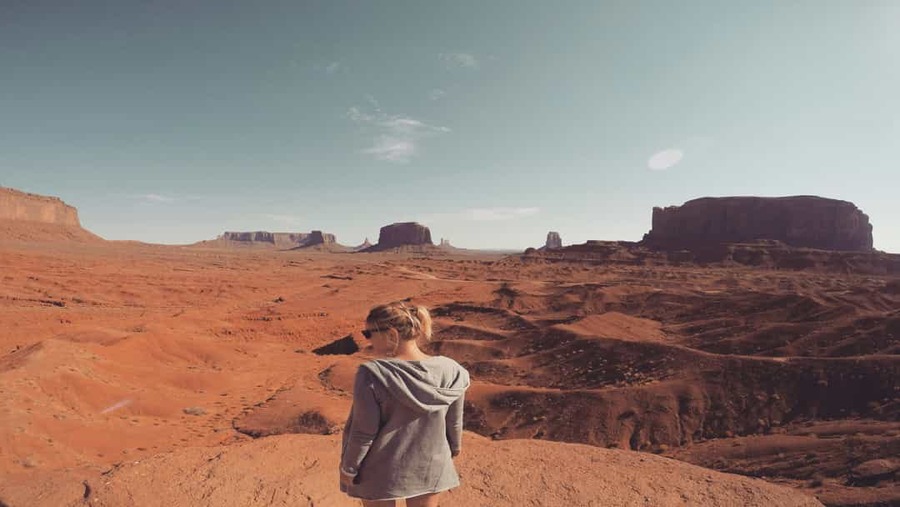 Vrouw met zonnebril staat op een rotsrand en kijkt uit over de rode buttes en mesa's van Monument Valley