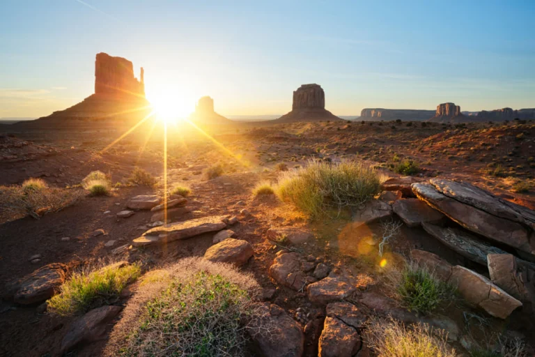 Opkomende zon tussen de karakteristieke rode rotstorens van Monument Valley met droge struiken en stenen op de voorgrond