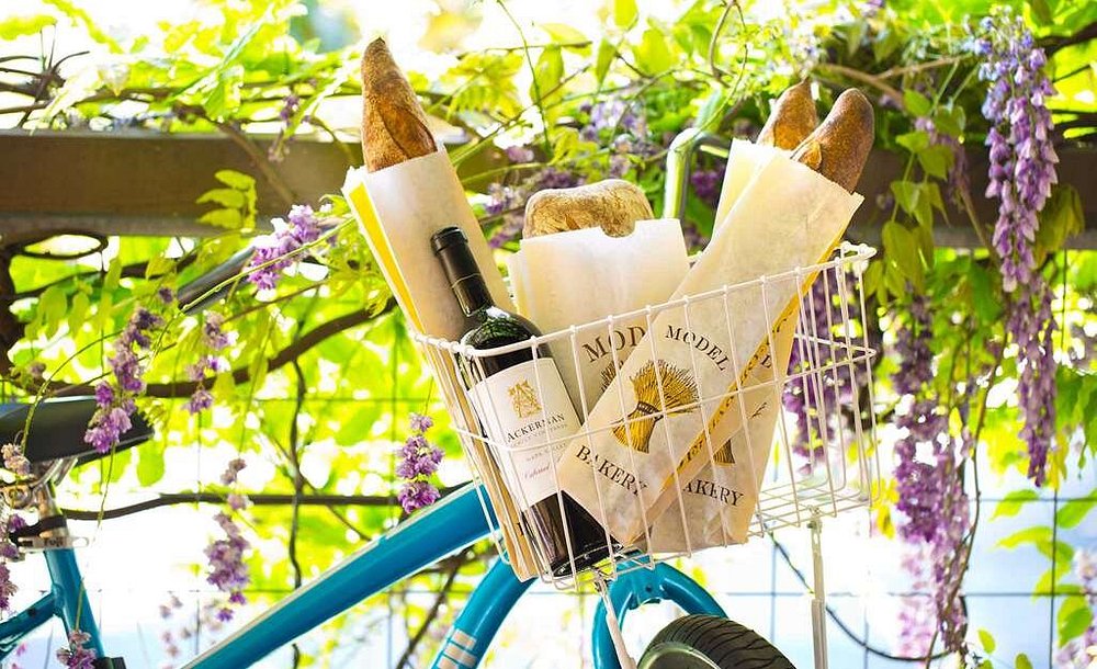 Witte fietsmand gevuld met stokbrood van een lokale bakkerij en een fles rode wijn hangt aan een fiets tussen paarse blauweregenbloesem