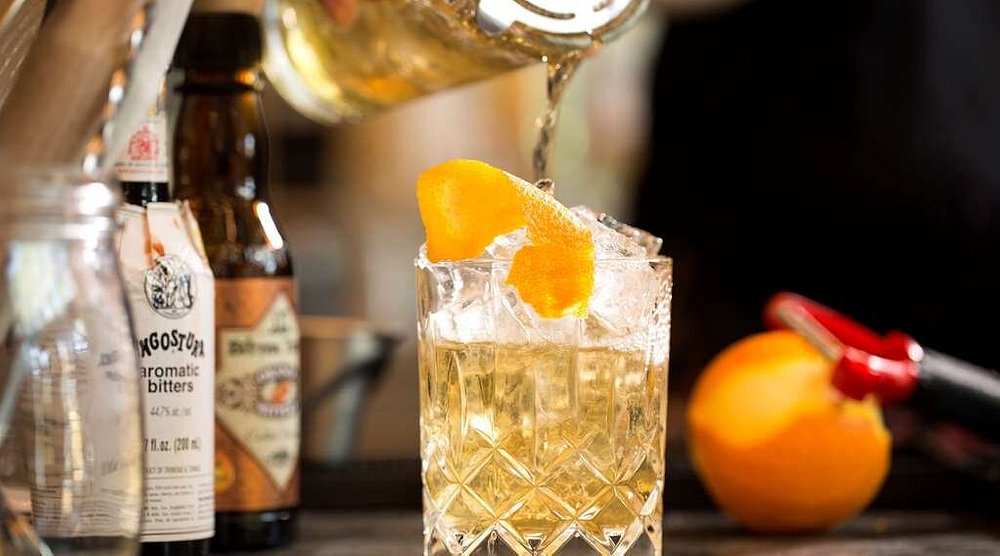 Amberkleurige cocktail met ijsblokken en een gedraaide sinaasappelschil wordt ingeschonken naast flesjes bitters op een bar