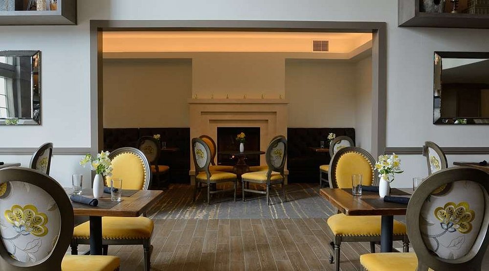 Ingerichte eetzaal met ronde tafels, gele stoffen stoelen en een marmeren open haard met kaarsen op de schoorsteenmantel