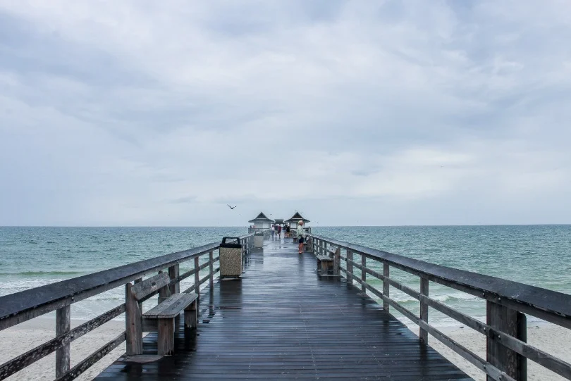 Een lange houten pier met bankjes loopt over het water bij Naples onder een bewolkte lucht