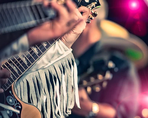 Close-up van een hand op de hals van een akoestische gitaar met franjejasje tijdens een muziekoptreden met roze podiumlicht op de achtergrond