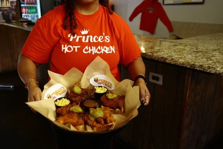 Een medewerkster in een oranje Prince's Hot Chicken shirt houdt een dienblad met gebakken kippenstukken en dipsausjes vast in Nashville