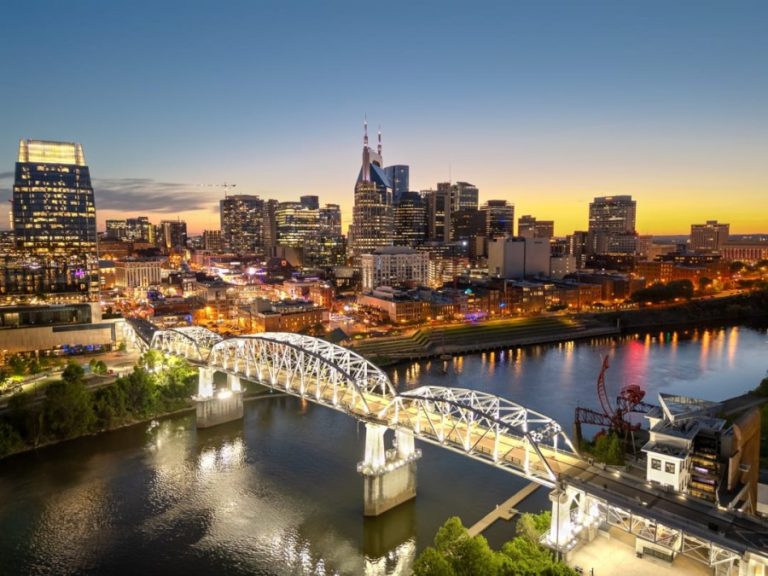 Verlichte skyline van Nashville met een witte voetgangersbrug over de rivier bij zonsondergang
