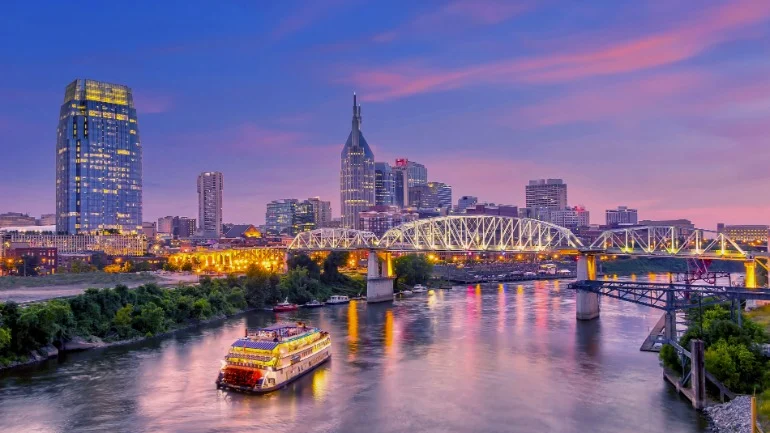 De skyline van Nashville met verlichte wolkenkrabbers en een brug over de rivier terwijl een rivierboot voorbij vaart bij zonsondergang