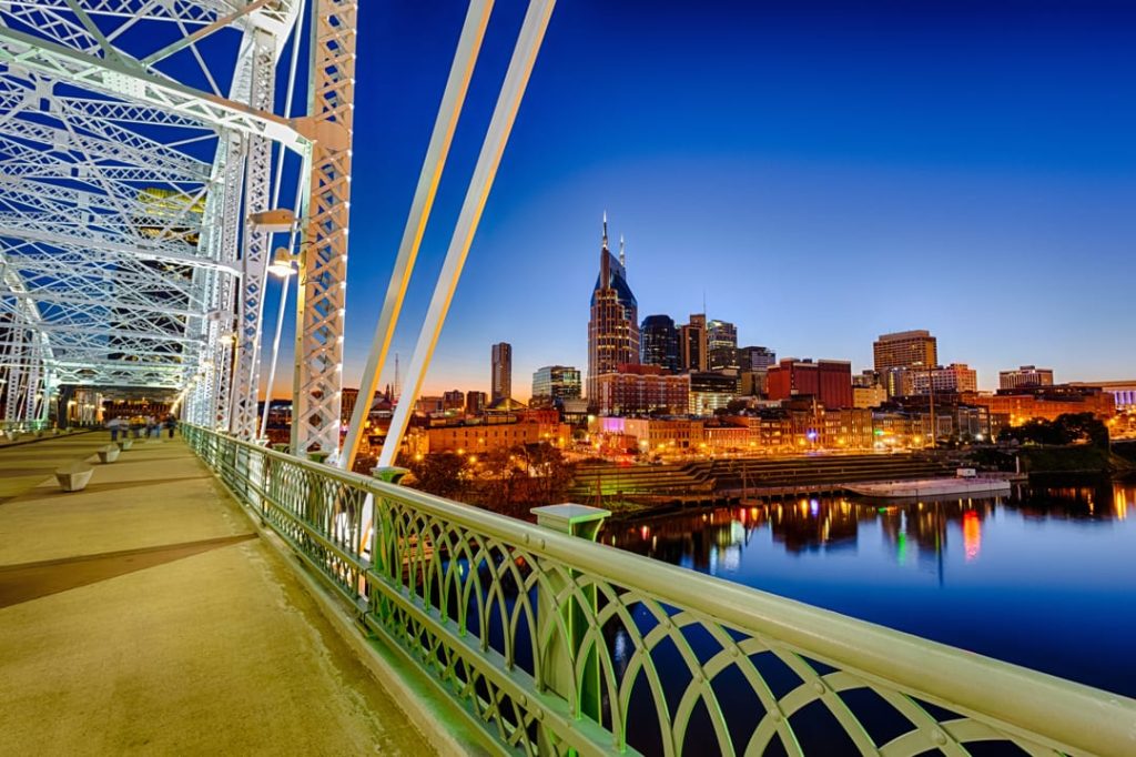 Verlichte stalen voetgangersbrug over de Cumberland rivier met de skyline van Nashville en de torenspitsen van het AT&T gebouw op de achtergrond bij nacht