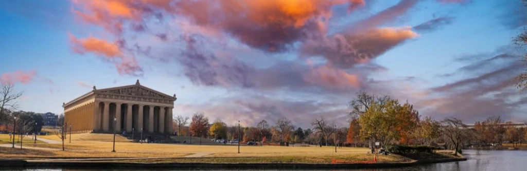 De replica van het Griekse Parthenon staat tussen bomen in een park terwijl de wolken oranje kleuren bij zonsondergang