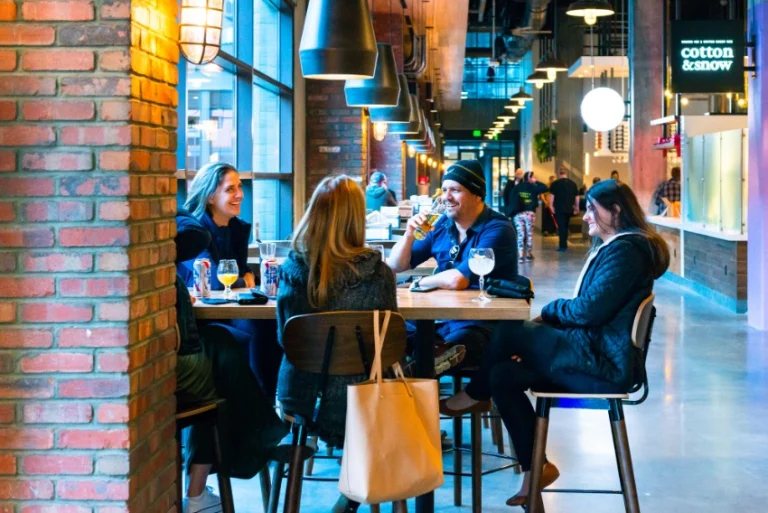 Vier vrienden zitten aan een houten statafel te drinken en te praten in een sfeervolle foodhall in Nashville met bakstenen muren en hanglampen