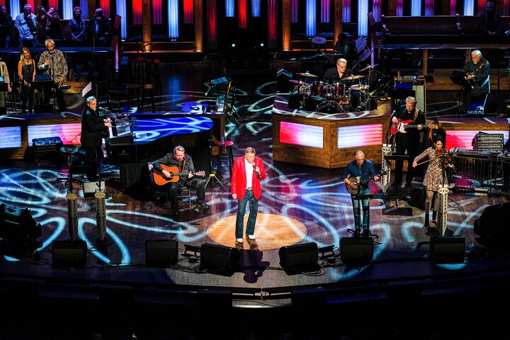 Zanger in rood jasje staat centraal op het podium van de Grand Ole Opry omringd door begeleidende muzikanten