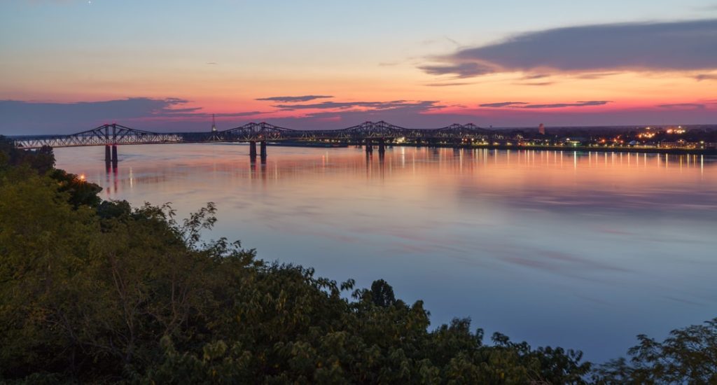 Roze en oranje zonsondergang boven de brede Mississippi rivier met twee stalen bruggen in de verte gezien vanaf een beboste oever