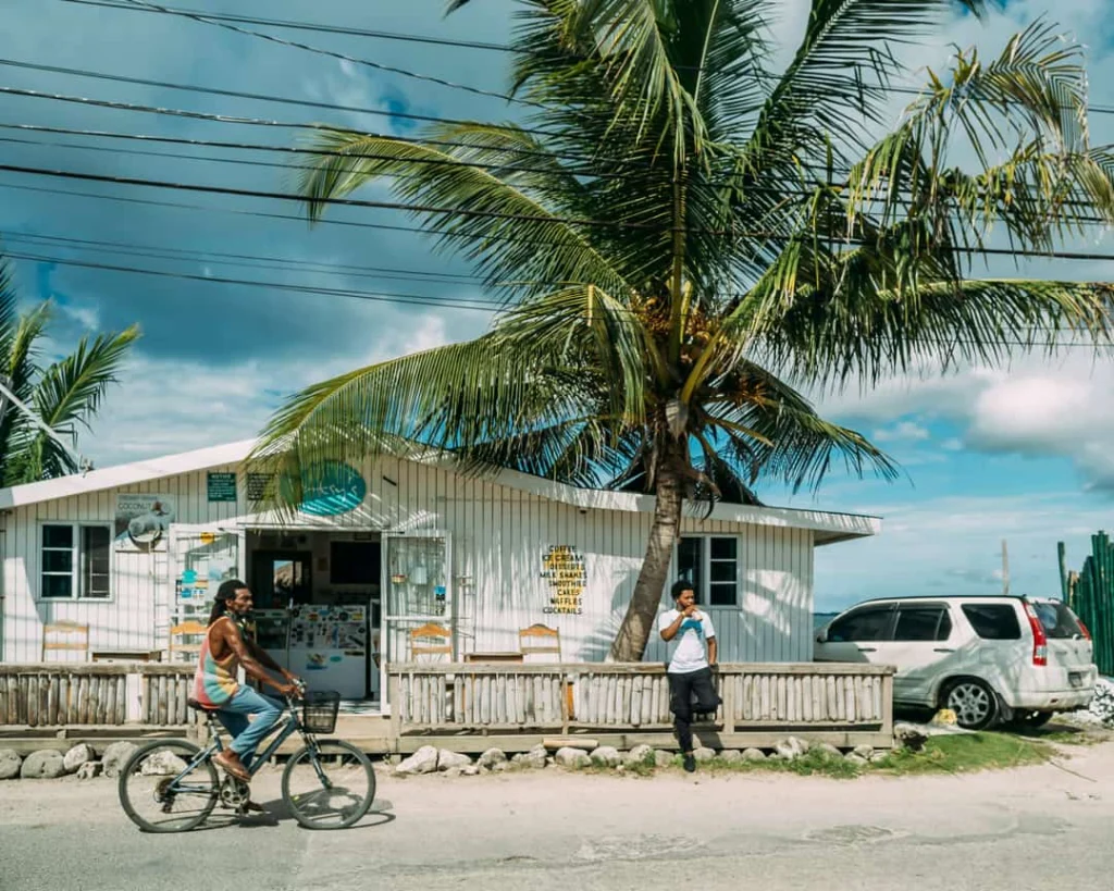 Een man fietst langs een wit houten strandhuisje met een grote palmboom ervoor terwijl iemand op de veranda staat in Negril