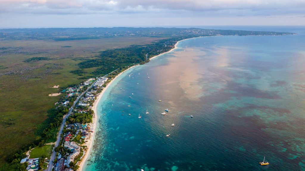 Luchtfoto van het lange witte strand van Negril met turquoise water, aangemeerde bootjes en groene kustvegetatie
