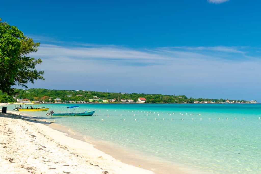 Wit zandstrand in Negril met twee gekleurde bootjes aan de waterlijn en turquoise zee onder een palmboom