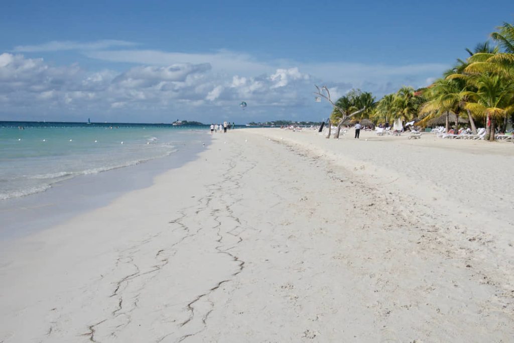 Wit zandstrand in Negril met palmbomen, ligstoelen en wandelaars langs de waterlijn onder een bewolkte lucht