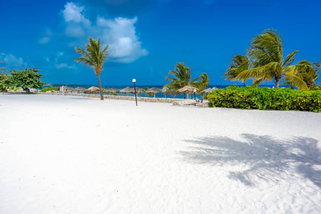 Leeg wit zandstrand in Negril met de lange schaduw van een palmboom en enkele strandhutjes op de achtergrond