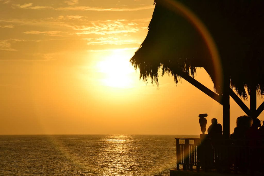 Silhouetten van mensen onder een rieten dak bij Rick's Cafe in Negril tijdens een gouden zonsondergang boven zee