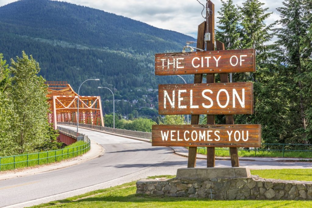 Houten welkomstbord met de tekst the city of Nelson welcomes you staat naast een weg met een oranje stalen brug
