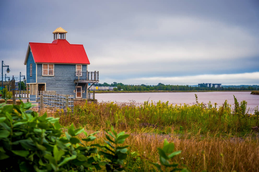 Een blauw houten huis met een opvallend rood dak en een torentje staat aan de oever van een rivier omringd door hoog gras