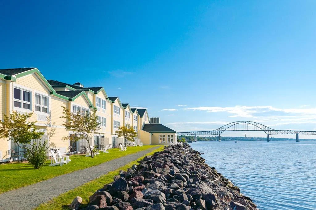 Een rij gele motelkamers met puntdaken ligt langs een rotsachtige waterkant, met een boogbrug op de achtergrond in New Brunswick