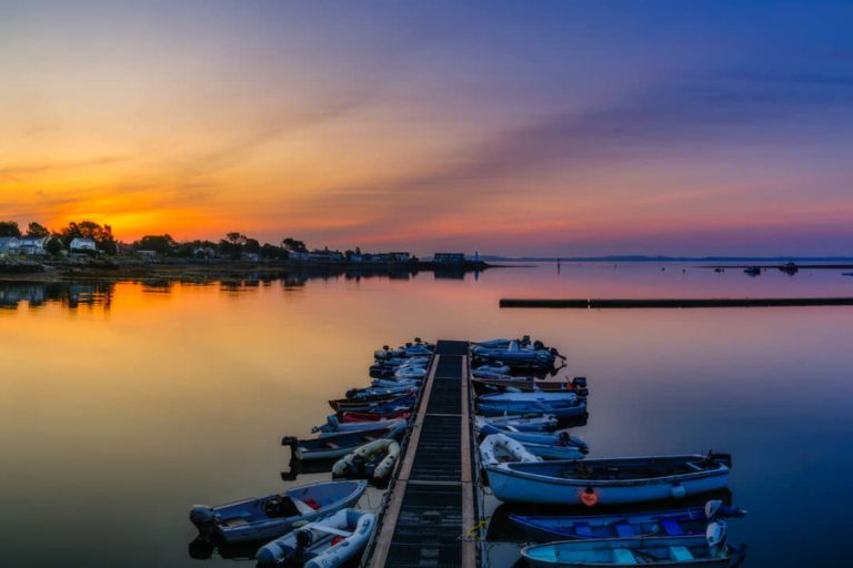 Kleine bootjes liggen afgemeerd aan een steiger in een jachthaven, met een oranje zonsondergang boven het water in New Brunswick