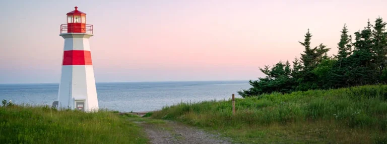 Een kleine vuurtoren met rode en witte banden staat op een grasveld aan zee bij zonsondergang in New Brunswick