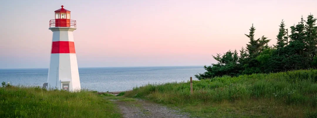 Een kleine vuurtoren met rode en witte banden staat op een grasveld aan zee bij zonsondergang in New Brunswick