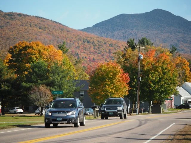 Auto's rijden over een dorpsstraat met felgekleurde herfstbomen en een berg op de achtergrond in New England