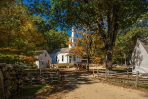 Wit houten kerkje met torenspits tussen herfstbomen, met een paard en wagen op een zandpad in een historisch dorp in New England