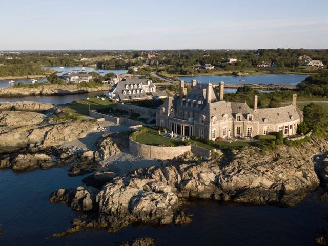 Groot stenen landhuis met torentjes op een rotsachtige kaap omringd door water en groene tuinen in New England