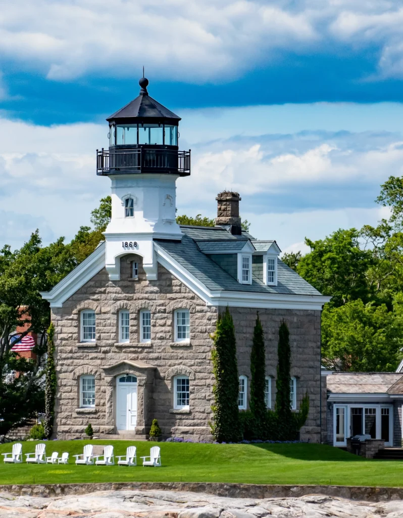 Witte vuurtoren met zwarte lantaarnkamer naast een stenen huis met tuinstoelen op het gras aan het water in New England