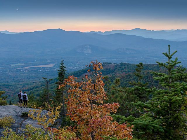 Twee wandelaars staan op een rotsig uitkijkpunt met een dal en bergketens in de avondschemering in New England