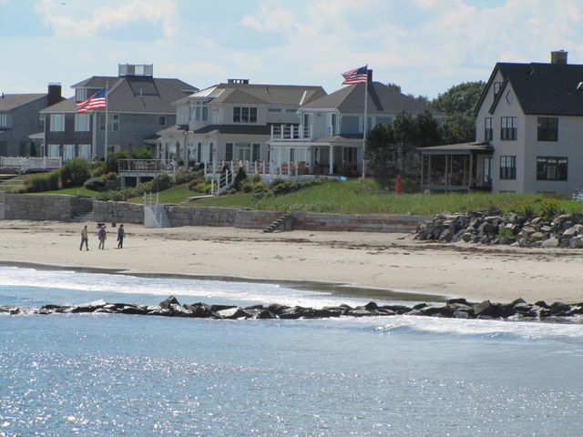 Grote houten strandhuizen met Amerikaanse vlaggen achter een lage stenen muur, met wandelaars op het zandstrand en een golfbreker van rotsen in New Hampshire