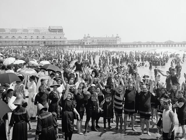 Zwart-witfoto uit het verleden van een grote groep badgasten met parasols op het strand voor een pier met theatergebouwen in New Jersey