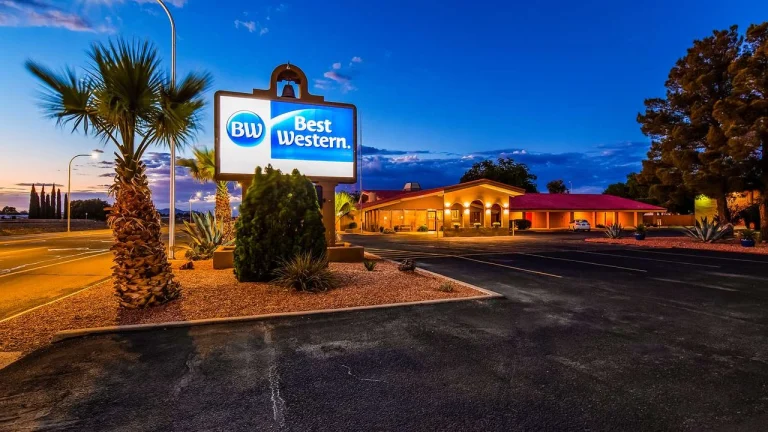 Verlicht bord van Best Western naast een laag hotelgebouw met rood dak, palmboom en cactussen bij het invallen van de avond
