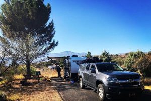 Pick-uptruck met caravan geparkeerd op een camping onder een pijnboom, met een picknicktafel en bergen op de achtergrond