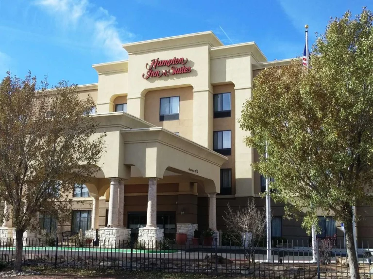 Beige hotelgebouw van Hampton Inn and Suites met een overkapte ingang en bomen ervoor onder een blauwe hemel