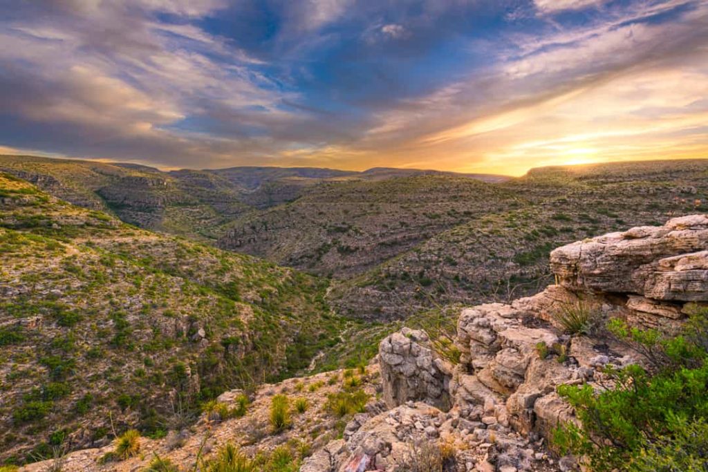 Uitgestrekt begroeid ravijn met rotsachtige kliffen op de voorgrond onder een dramatische, gekleurde avondlucht in New Mexico