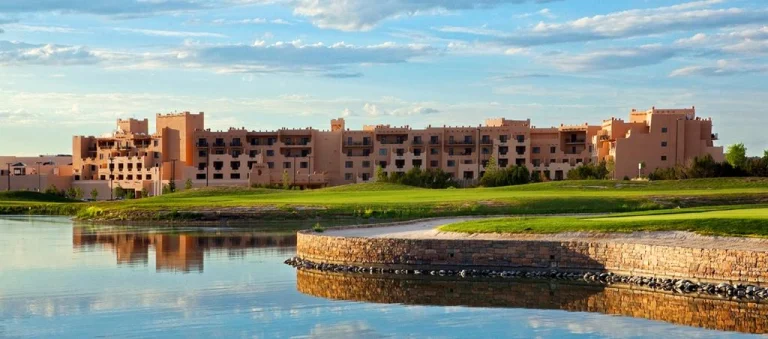 Laag terracottakleurig resortgebouw in pueblostijl met een golfbaan en kunstmatig meer ervoor onder een lichtbewolkte hemel