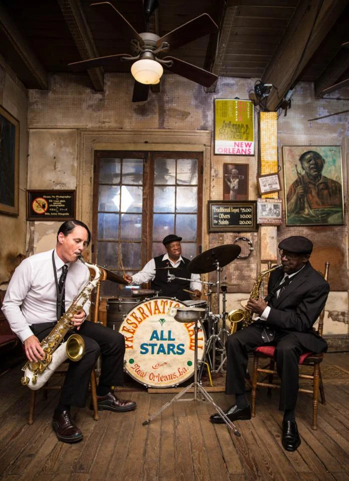 Drie muzikanten met saxofoons en drum spelen in het intieme houten interieur van Preservation Hall in New Orleans, met een grote bassdrum op de voorgrond