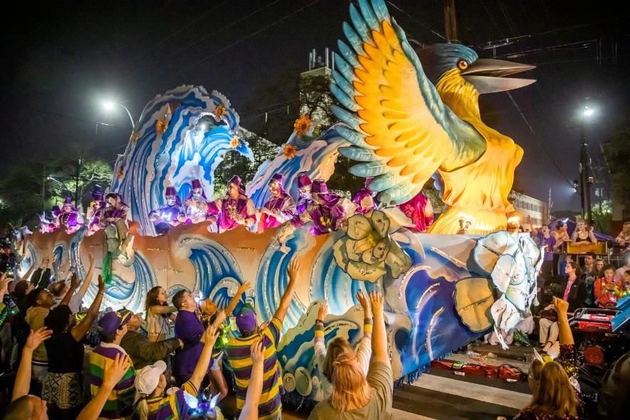 Bij nacht rijdt een grote praalwagen met een gouden vogel en golvende decoraties door de straten van New Orleans, terwijl feestgangers naar boven reiken tijdens de Mardi Gras parade