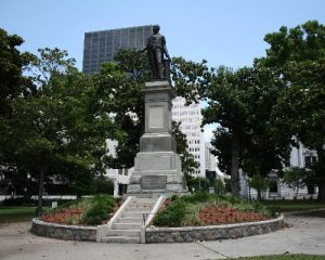 Bronzen standbeeld van een man op een hoog voetstuk in een park met bloemperken, bomen en moderne gebouwen op de achtergrond in New Orleans